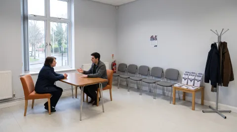 Zwei Personen sitzen an einem schlichten Klapptisch im hellen Foyer einer Beratungsstelle, an der Wand gegenüber steht eine Reihe leerer Wartestühle