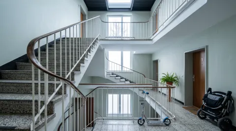 Blick nach oben durch ein Treppenhaus mit Terrazzostufen, auf jedem Podest liegt eine Wohnungstür, im Erdgeschoss stehen ein Kinderroller und ein zusammengeklappter Kinderwagen