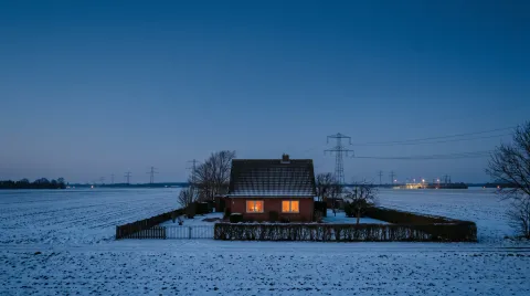 Wohnhaus mit warm erleuchteten Fenstern am Feldrand in der winterlichen Abenddämmerung, dahinter eine Hochspannungsleitung am Horizont.