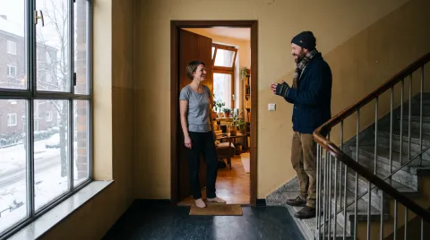 Zwei Nachbarn im Treppenhaus eines Mehrfamilienhauses: Sie steht im T-Shirt in ihrer Wohnungstür, er am Fuß der Treppe mit Mütze, Schal und Handschuhen.